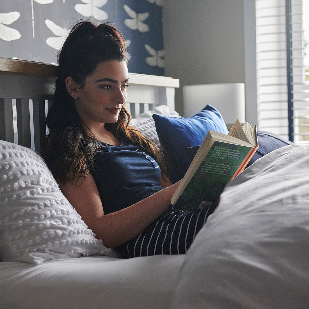 Woman reading a book in bed with white bedding and blue pillows.