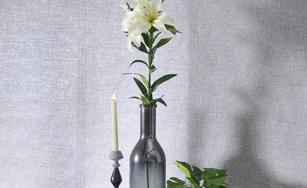 Decorative arrangement with a bottle vase holding a white flower against a gray textured wall.