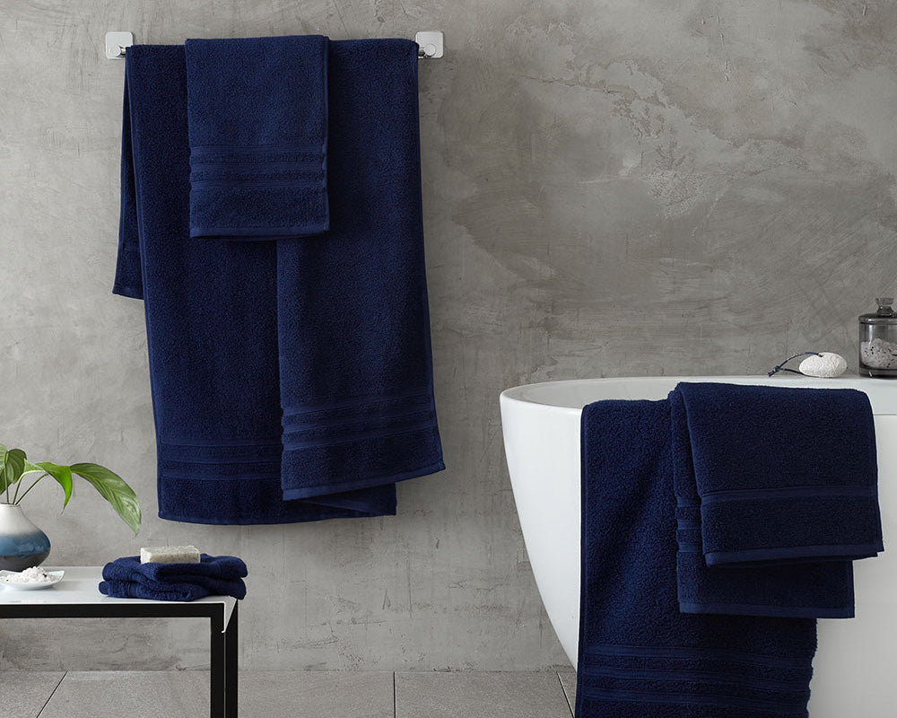 Navy blue towels hanging on a towel rack and draped over a white bathtub against a grey tiled wall.