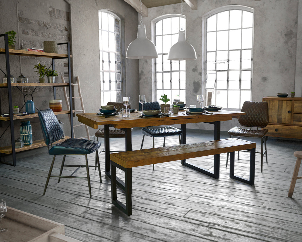 Dining area with wooden table, chairs, and bench in a room with large windows and industrial decor.