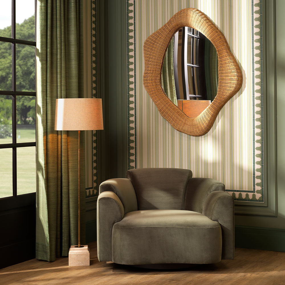 Grey armchair in a room with a wicker mirror, lamp, and striped wallpaper.