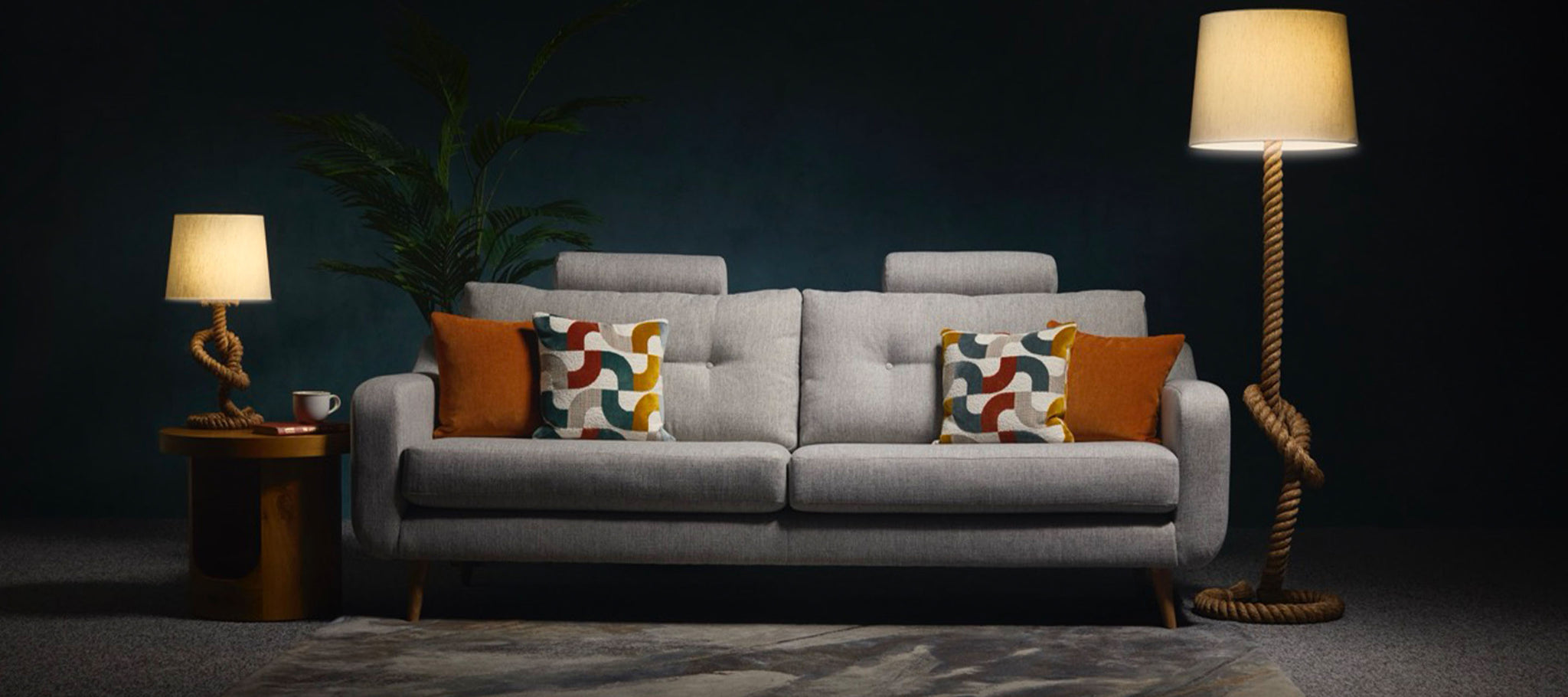 Grey sofa with colourful cushions in a dimly lit room with lamps and a plant.