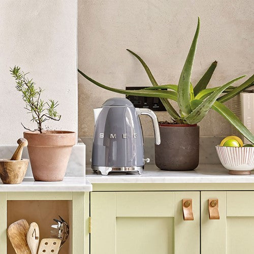 Gray Smeg kettle on a kitchen counter with potted plants and a bowl of fruit.