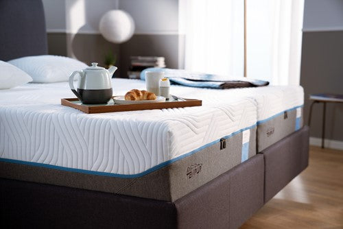 Bed with Tempur mattress and brown frame, tray with tea set and croissant on a wooden floor.