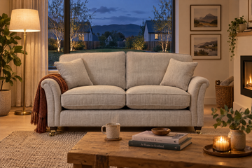 A light beige upholstered loveseat sofa in a cozy living room setting, styled with matching throw pillows, a brown throw blanket, a wooden coffee table, and a warm fireplace in the background.
