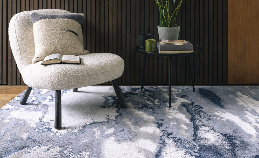 Beige armchair with a pillow on a textured blue and white rug in a room with wooden walls.
