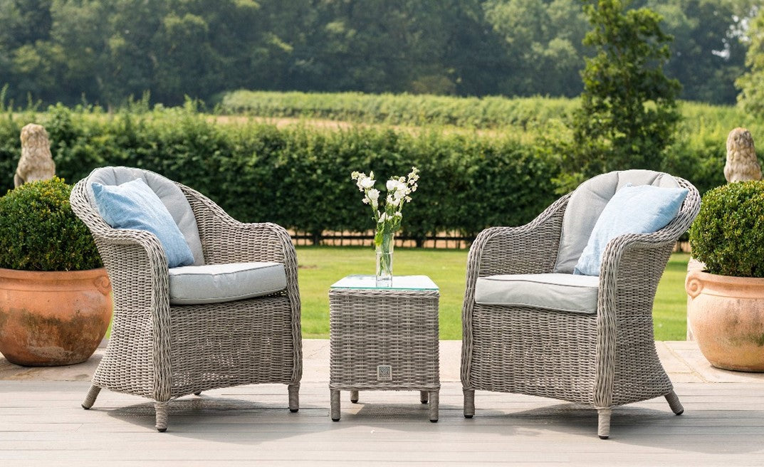 Two rattan outdoor chairs with cushions and a rattan small table on a patio with greenery in the background.