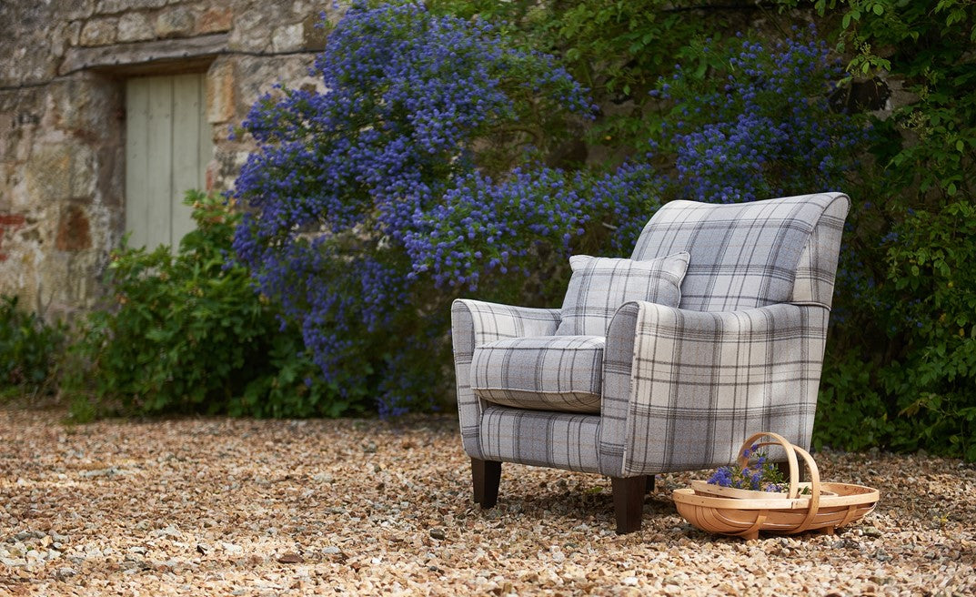 Plaid armchair in a garden setting with a stone building and blue flowers in the background.