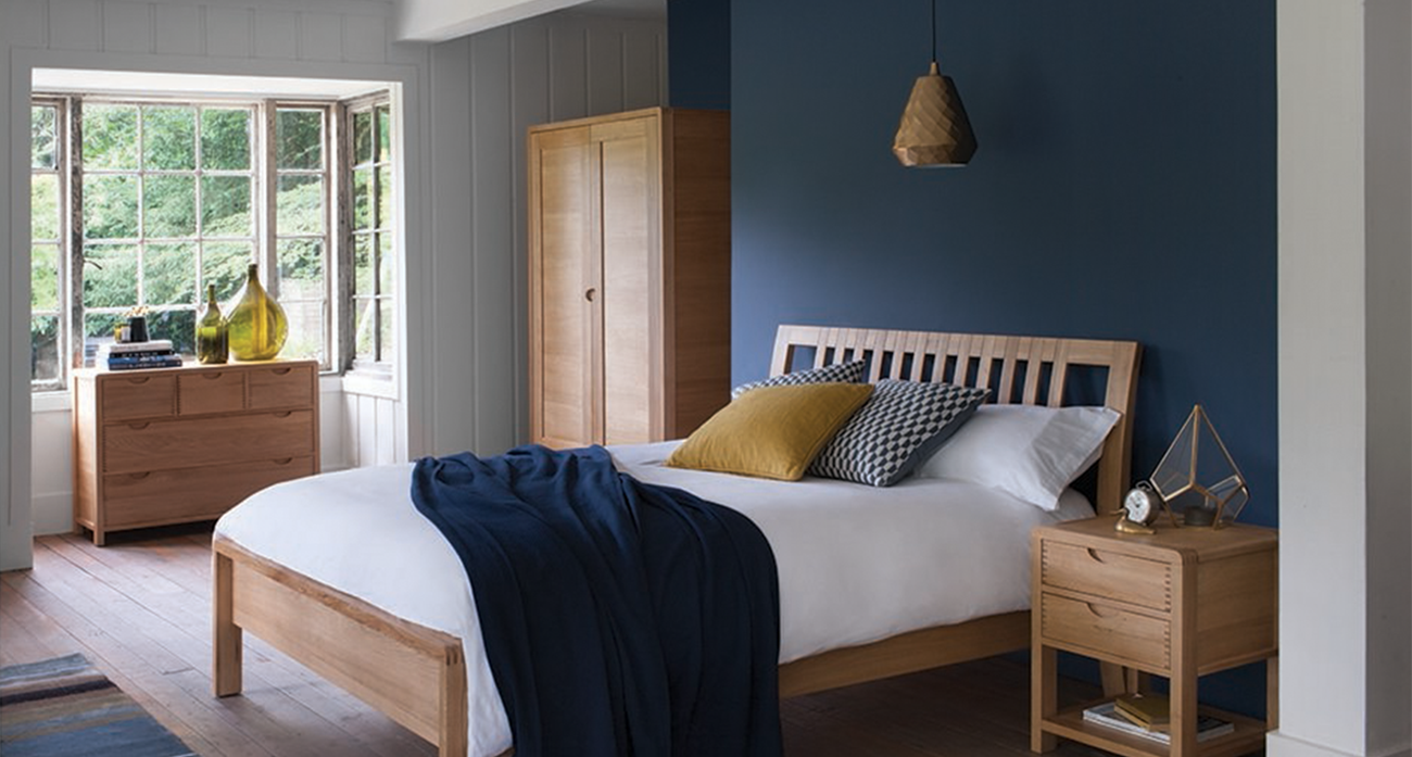 Ercol Bedroom furniture with wooden bed frame, bedside table, and wardrobe against a blue wall.